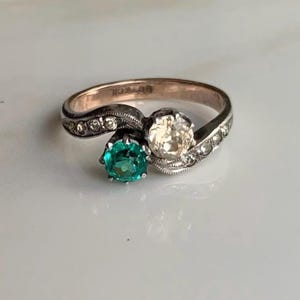 Op de afbeelding: Een zilveren en roségouden ring met een groene smaragd en een witte diamant. De ring is bezet met kleine diamanten rond de band.