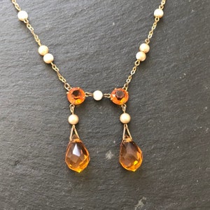 May include: A gold necklace with a delicate chain and small pearl accents. The necklace features a pendant with two teardrop-shaped amber stones and two round amber stones.