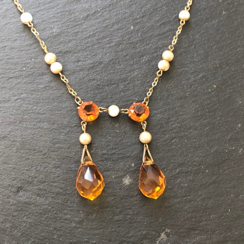 May include: A gold necklace with a delicate chain and small pearl accents. The necklace features a pendant with two teardrop-shaped amber stones and two round amber stones.