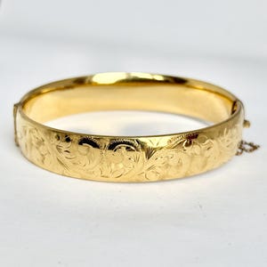 May include: A gold-coloured bangle bracelet with an engraved floral pattern. The hinged bracelet has a safety chain and a shiny, polished finish. The design features detailed, repeating floral and leaf motifs.