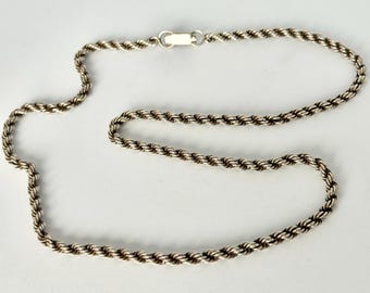 May include: A silver-toned rope chain necklace with a lobster clasp. The chain is made of twisted strands, creating a textured, rope-like appearance. The necklace is displayed against a plain white background.