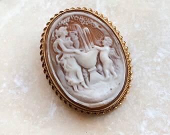 May include: A gold-toned oval cameo brooch with a white cameo depicting three figures in a garden setting. The figures are carved in relief and appear to be interacting with a fountain.