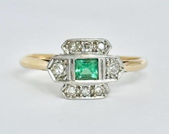 May include: A gold ring featuring a central square-cut green gemstone set in a rectangular white metal head. The setting is accented with small round clear stones arranged in rows above and below the center stone and on the sides of the frame.