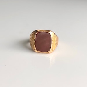 May include: A gold ring featuring a square, reddish-brown stone set in the centre. The ring is decorated with intricate engravings around the stone.