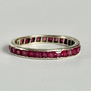 Peut inclure: Une bague en argent ornée d'une rangée continue de pierres précieuses rouge foncé taillées en carré. Le métal de la bague a un aspect légèrement vieilli, et les pierres sont serties de près. Un bijou.
