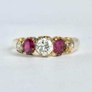Op de afbeelding: Een gouden ring met een centrale briljant geslepen diamant, geflankeerd door twee ronde rode robijnen en twee kleinere diamanten aan de buitenranden. De stenen zijn gezet in een gepolijste gouden band met een glad, afgerond profiel.