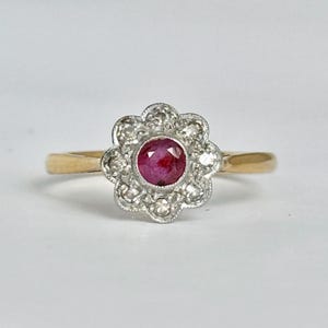 May include: A gold ring featuring a central round-cut red ruby surrounded by a floral-shaped cluster of small, clear, round-cut diamonds set in a silver-toned metal setting. The band is a smooth, polished yellow gold metal.
