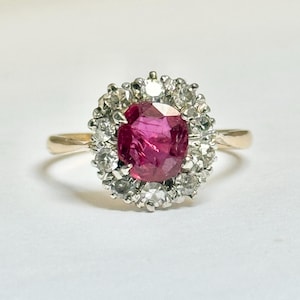 Può includere: Anello d'oro con un rubino centrale ovale, circondato da un alone di piccole pietre preziose chiare. Il rubino è di un rosso intenso e la montatura è in oro.