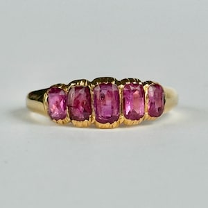 May include: A gold ring with five rectangular pink gemstones. The gemstones are set in a row, with a gold band. The ring is displayed against a white background, highlighting the ring's design and colour.