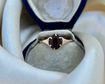 Könnte beinhalten: Ein goldener Ring mit einem dunkelroten ovalen Edelstein, der in einem schlichten und eleganten Design gefasst ist. Der Ring wird in einer blauen Samt-Ringbox präsentiert.