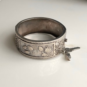 May include: A silver hinged bangle bracelet with an engraved floral design and a dangling key charm. The bracelet is marked with the number "925" on the inside.