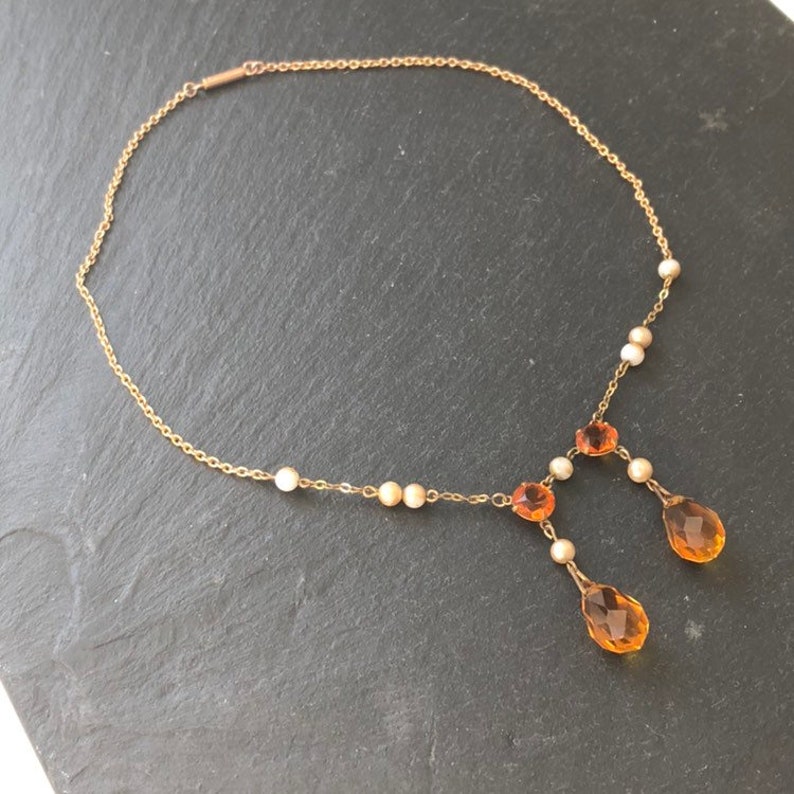 May include: A gold chain necklace with a teardrop-shaped pendant. The pendant is made of amber glass and has a faceted design. The necklace also features small white and gold beads.