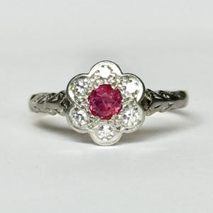 Könnte beinhalten: Ein silberfarbener Ring mit einem zentralen, rund geschliffenen, rötlich-pinken Edelstein, der von sechs kleineren, klaren, rund geschliffenen Steinen in einer blütenförmigen Anordnung umgeben ist. Die Metallschiene zeigt ein eingraviertes Muster.