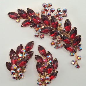 May include: A jewelry set consisting of a brooch and a pair of matching earrings. Each piece features deep red marquise-cut rhinestones arranged in a leaf-like pattern, accented with smaller, iridescent, round-cut crystals set in gold-tone metal.