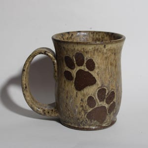 May include: A ceramic mug with a tan and brown mottled glaze. Two dark brown paw prints are featured on the front of the mug. The mug has a rounded handle and a slightly flared rim. This mug is perfect for coffee or tea.