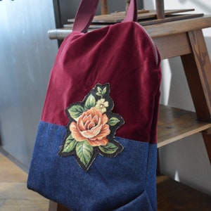 May include: A burgundy velvet tote bag with a denim bottom and a floral embroidered patch. The bag has a zipper closure and a long strap.