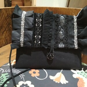 Puede incluir: Un bolso de mano negro de tela con ribete de cuentas plateadas y negras. El bolso tiene cierre de solapa y una correa corta.