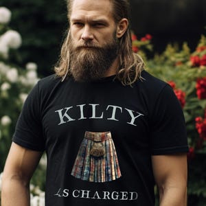 May include: Black t-shirt with the text "KILTY" above a graphic of a kilt and sporran, with the words "AS CHARGED" below. The shirt is worn by a person with long blond hair and a beard.