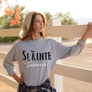 May include: A gray long-sleeve sweatshirt with the text "Sl&agrave;inte Sassenach!" printed in black and white. The model is wearing the sweatshirt with a white beanie and a plaid shirt tied around their waist.