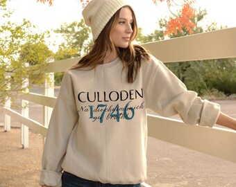 May include: A beige sweatshirt with the text "CULLODEN" and "Na diephyl ma nick 1746" printed on the front. The model is wearing a white beanie and blue jeans.