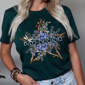 May include: A dark green t-shirt with a five-pointed star design featuring blueberries, twigs, and leaves. The text "Ban-Ortuidh" is printed in a stylized font inside the star.