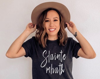 May include: A dark gray t-shirt with the words "Slainte mhath" in white script. The person in the image is wearing a brown hat and blue jeans. The shirt is a casual style.