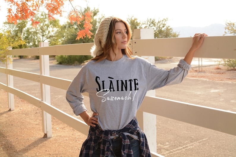 May include: A gray long-sleeve sweatshirt with the text "Sl&agrave;inte Sassenach!" printed in black and white. The model is wearing the sweatshirt with a white beanie and a plaid shirt tied around their waist.