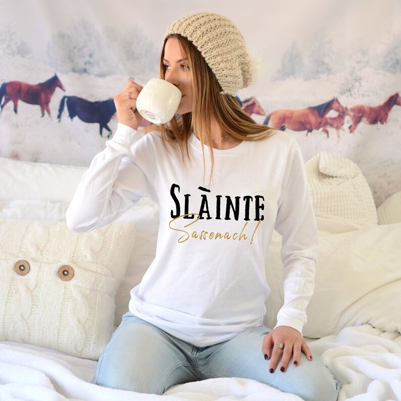 May include: White long-sleeve shirt with black text that reads "Sl&aacute;inte Sassenach!" in a vintage style font. The text is in gold and black.