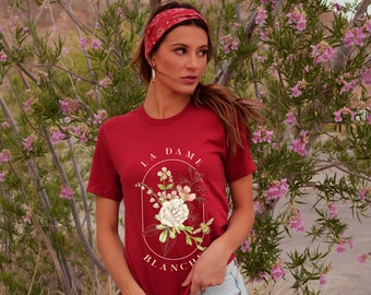 May include: A red t-shirt with a floral design and the text "La Dame Blanche" in a circle.