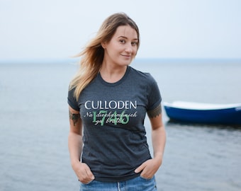 May include: A woman wearing a dark gray t-shirt with the text "CULLODEN Na  deo  ch  am  b  nich  gu  br  ath  1746" printed on it. She is standing on a beach with a boat in the background.