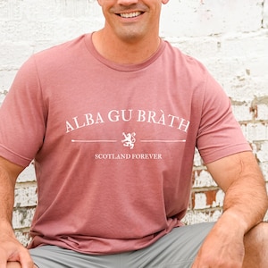 Puede incluir: Una camiseta de color rosa pálido con un diseño gráfico blanco que dice "Alba Gu Bràth" con un emblema de león y el texto "Scotland Forever".