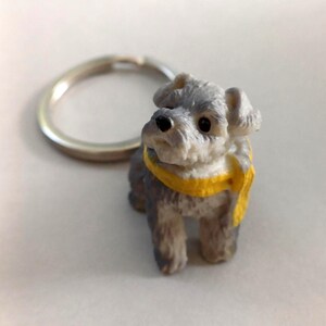 Puede incluir: Un llavero de perro Schnauzer miniatura con un anillo plateado. La figura del perro es gris y blanca con una bufanda amarilla. Un accesorio encantador.
