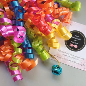 May include: A decorative gift bow featuring multi-coloured curly ribbons in shades of pink, orange, yellow, blue, and lime green. A small metallic blue bell hangs from a gold string. A square white card displays the text from Bowzandgiftz on Etsy.com.
