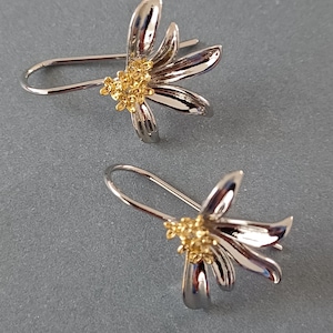 May include: A pair of silver-tone flower earrings with gold-coloured centres. Each earring features a curved hook and a stylised floral design with multiple petals. The earrings are displayed on a grey background.