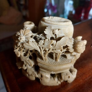 May include: A beige stone carving featuring a central vessel surrounded by intricate relief work of chrysanthemum flowers and foliage. The sculpture displays detailed petals, leaves, and a small cylindrical opening, all crafted from a single piece of material.