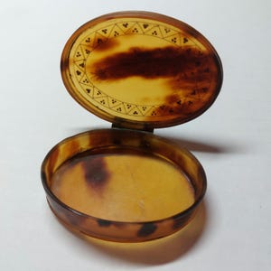 May include: A small, round, tortoiseshell trinket box with a hinged lid. The lid is decorated with a geometric pattern and a small insect design.