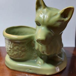 May include: A green ceramic planter shaped like a dog. The dog is sitting with its head turned to the side and its tail curled around its legs. The planter has a small pot on its back for planting.