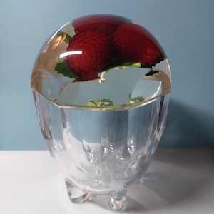 May include: A clear glass decorative bowl with a domed lid, containing three red raspberries and green leaves. The bowl has a faceted design and sits on three small feet. The raspberries are encased in a clear resin.
