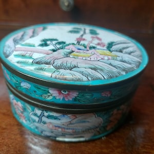 May include: An oval trinket box with a detailed landscape scene on the lid. The box has a teal base with floral designs and a brass rim. The lid depicts a serene landscape with mountains, trees, and buildings.