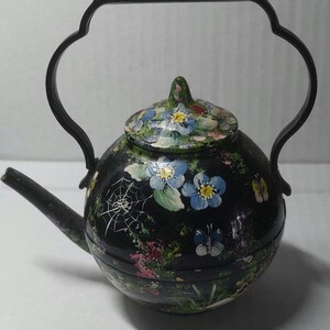 May include: A round black teapot featuring a hand-painted design of blue flowers, green foliage, small butterflies, and a white spider web. The teapot has a black lid with a matching floral pattern and a tall, arched black metal handle.