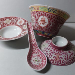 May include: A collection of red and white ceramic tableware with an Asian design. The set includes a bowl, a small dish, a spoon, and a cup with a saucer. Each item is decorated with a floral pattern and a Chinese character.