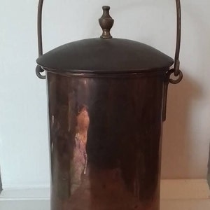 May include: A vintage copper bucket with a lid and a handle. The cylindrical bucket has a dark brown patina, with a matching lid and a curved handle. The handle is attached to the bucket with metal rings. The lid is topped with a small, decorative finial.