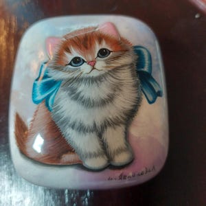May include: A hand-painted, rectangular trinket box featuring a fluffy ginger, white, and grey kitten with blue eyes and a blue bow. The kitten is centered on a pearlescent background with a pink and blue gradient.