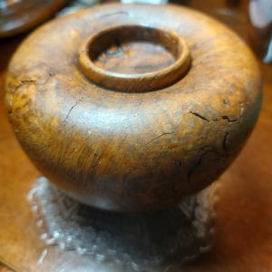 May include: A wooden bowl with a circular design and a raised, ring-shaped centre. The bowl has a natural wood grain pattern and a warm, brown colour. The bowl is sitting on a clear, decorative base.