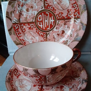 May include: A red and gold teacup and saucer set with a floral and geometric pattern. The saucer has a large central design with a red circle containing Chinese characters. The teacup has a white interior.