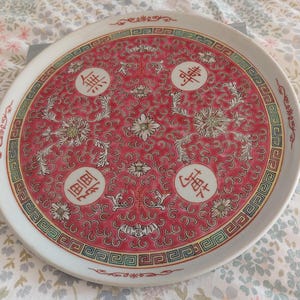 May include: A round, white ceramic plate with a red centre featuring floral and Chinese character designs. The plate has a decorative border with green, red, and gold accents. The plate is a decorative item.