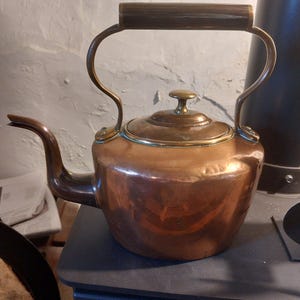 May include: A copper kettle with a wooden handle and brass detailing. The curved spout and brass lid knob are prominent features. This kettle, likely used for boiling water, has a classic design.