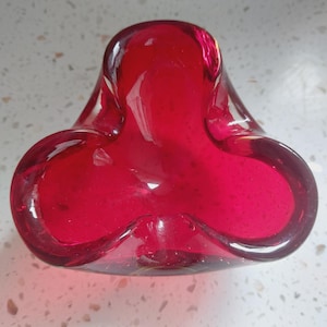 May include: A striking red glass bowl with a distinctive three-lobed form. The bowl has a smooth, glossy surface and an organic shape. Tiny air bubbles are visible within the glass. This decorative item is suitable for holding small objects.