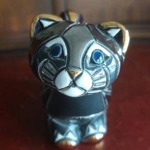 May include: A decorative cat figurine with a grey body, gold accents, and blue eyes. The cat has a white muzzle and a black collar. The figurine is made of a metallic material and has a detailed design.