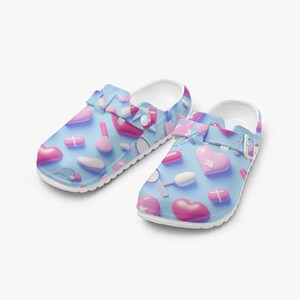 Op de afbeelding: Een paar blauwe klompen met een witte zool, versierd met roze harten, pillen en medische instrumenten. De verstelbare band heeft een vergelijkbaar patroon. De schoenen zijn ontworpen met een medisch thema.